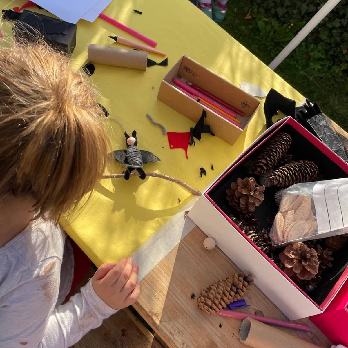 Blick von oben auf einen Basteltisch mit gelber Tischdecke, an dem ein Kind mit Naturmaterialien kleine Fledermaus-Figuren bastelt.