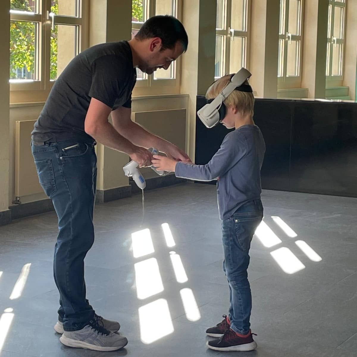 Mann und Junge stehen sich in einem Raum mit Fenstern gegenüber. Der Mann beugt sich leicht nach unten, um den Jungen, der eine VR-Brille trägt, zu unterstützen.