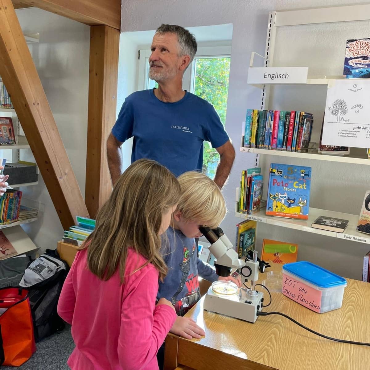 Zwei Kinder und ein Mann in einer Bibliothek. Eines der Kinder schaut in ein Mikroskop. 