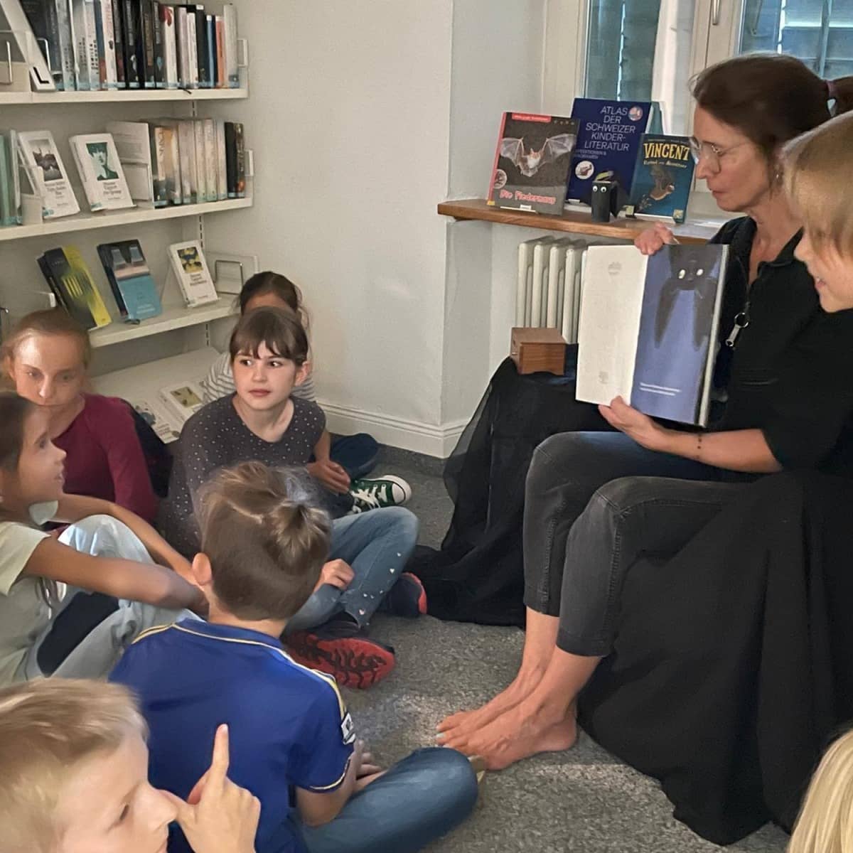 Kinder sitzen in einer Bibliothek am Boden und schauen zu einer Frau, die ihnen etwas auf dem Ipad präsentiert.