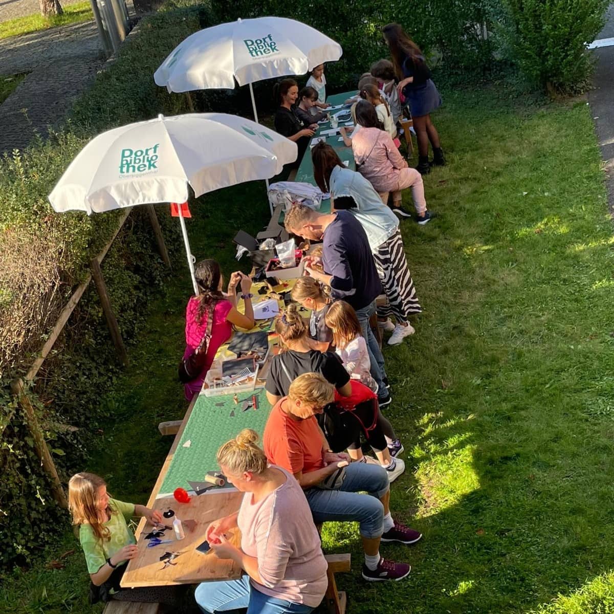 Blick von oben auf einen langen Basteltisch auf einer Rasenfläche. Am Tisch sitzen Erwachsene und Kinder unter Sonnenschirmen und basteln gemeinsam.