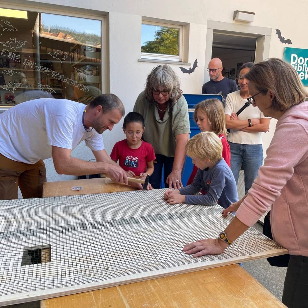 Kinder und Erwachsene stehen vor der Dorfbibliothek um einen Holztisch und werkeln. Auf dem Tisch liegt ein grosses Holzbrett mit kleinem viereckigem Loch und mit einem Gitter überspannt. Die Fassade der Dorfbibliothek ist mit Fledermäusen aus Papier geschmückt.