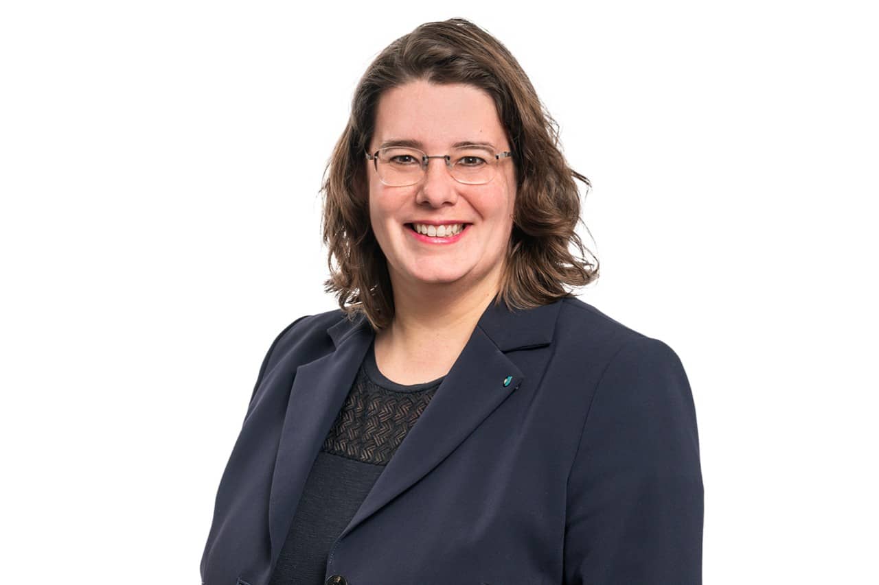 Eine Frau mittleren Alters mit schulterlangen, gewellten, braunen Haaren lächelt leicht seitlich in die Kamera. Sie trägt einen grauen Blazer.