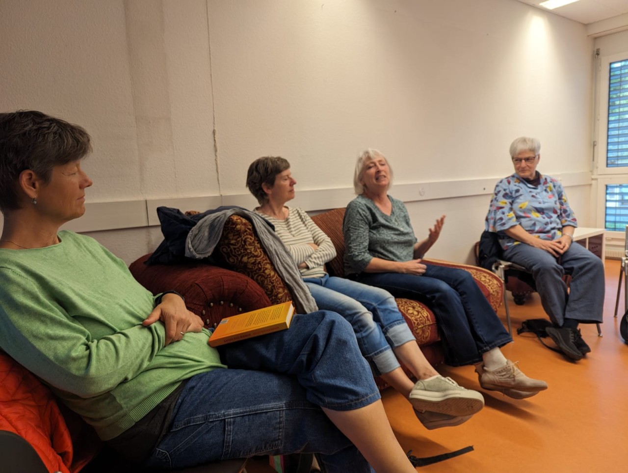 Ein Gruppe von Frauen mittleren Alters sitzen im Halbkreis auf Stühlen und diskutieren über ein Buch. Das Buch liegt auf dem Schoss der Frau im linken Rand des Bildes.