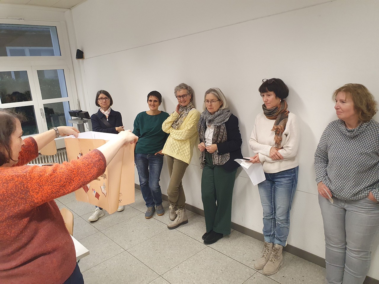 Das Foto zeigt eine Person in der rechten unteren Ecke, die eine Maquette einer Tasche in die Höhe hält. Ihr gegenüber stehen sechs Personen an einer Wand und betrachten diese Tasche gespannt.