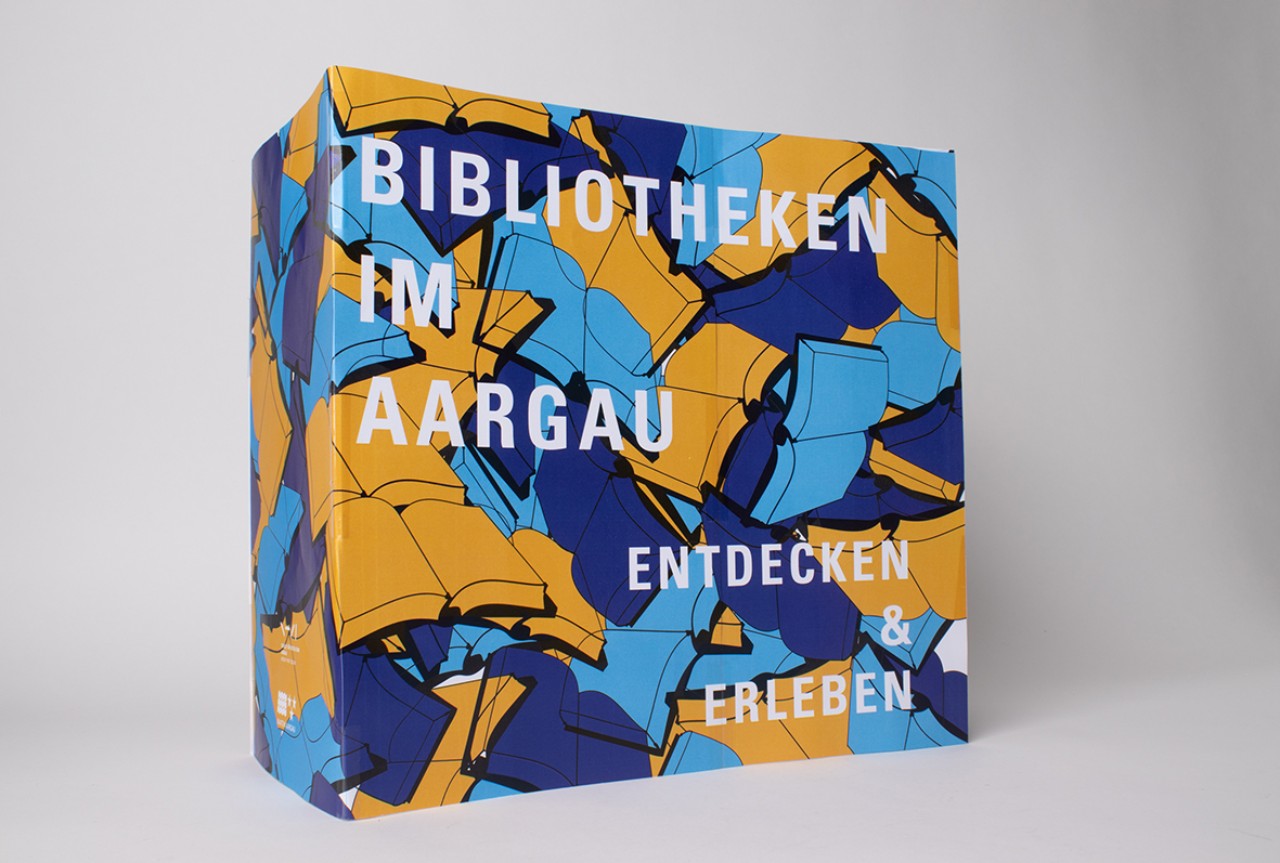 Die Maquette der Tasche aus Papier zeigt den Entwurf einer Schülerin. Das Sujet besteht aus wild übereinander geworfenen Büchern, die sich an vielen Ecken und Kanten überlappen. Farblich wechseln sich die aufgeschlagenen Büchern ab zwischen Orange, Dunkelblau und Cyan. 