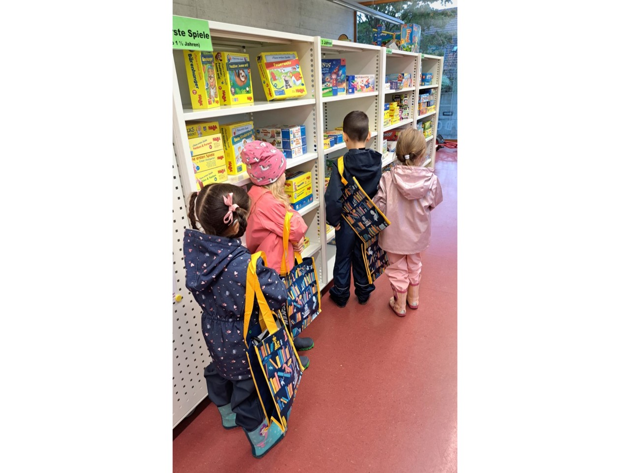 Vier Kinder stehen mit dem Rücken zur Kamera vor einem Regal mit Gesellschaftsspielen und tragen gestaltete Taschen um die Schultern. Die Taschen sind dunkelblau mit gelbem Rand und gelben Trägern und zeigen Figuren, die ein Bücherregal erkunden.