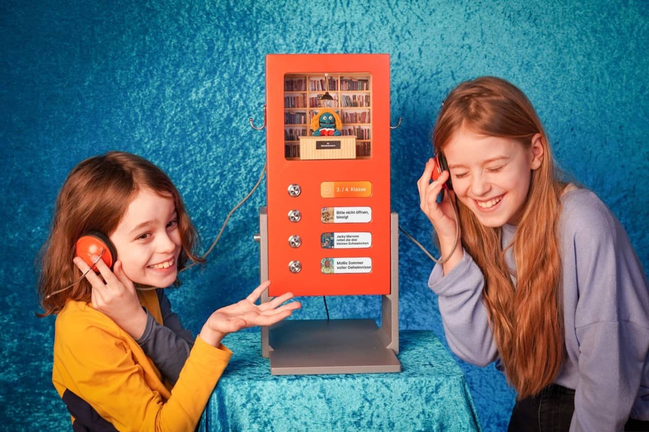 Zwei Kinder halten sich Kopfhörer ans Ohr und lachen in die Kamera. In der Mitte steht ein kleiner roter Automat mit Knöpfen, in dem eine kleine Bibliothek eingerichtet ist.