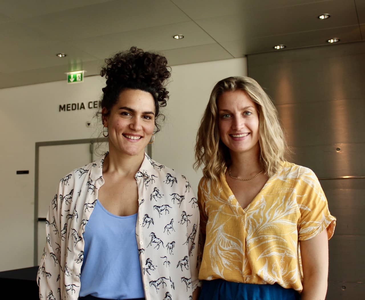 Ein Portraitbild zweier Frauen, die sich seitlich umarmen. Die Frau links hat dunkles, gelocktes Haar, das zu einem wilden Dutt hochgebunden wurde, sie recht hat schulterlanges, blondes Haar. Beide Frauen lächeln sympathisch in die Kamera.