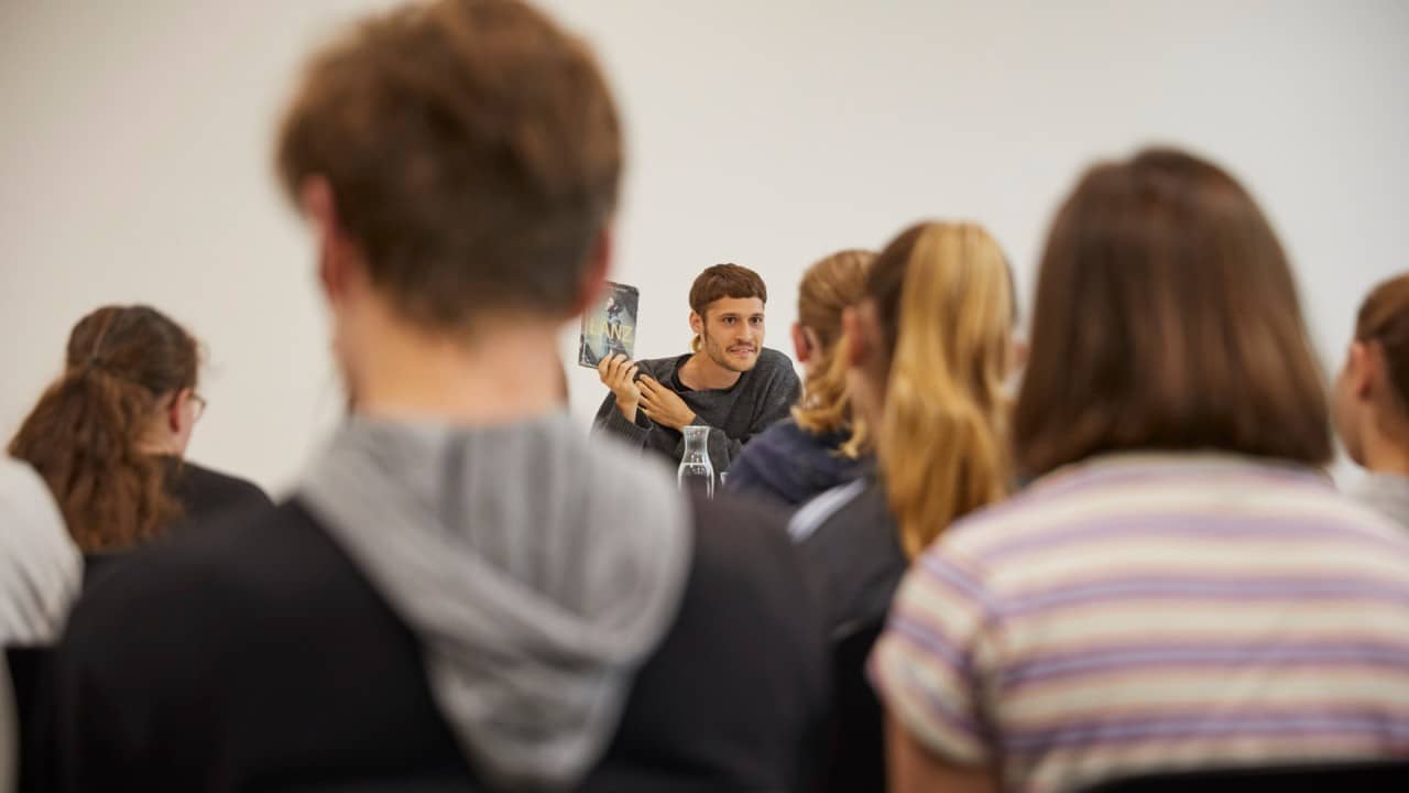 Autor Flurin Jecker sitzt bei einer Lesung vor einer Klasse und zeigt sein Buch.