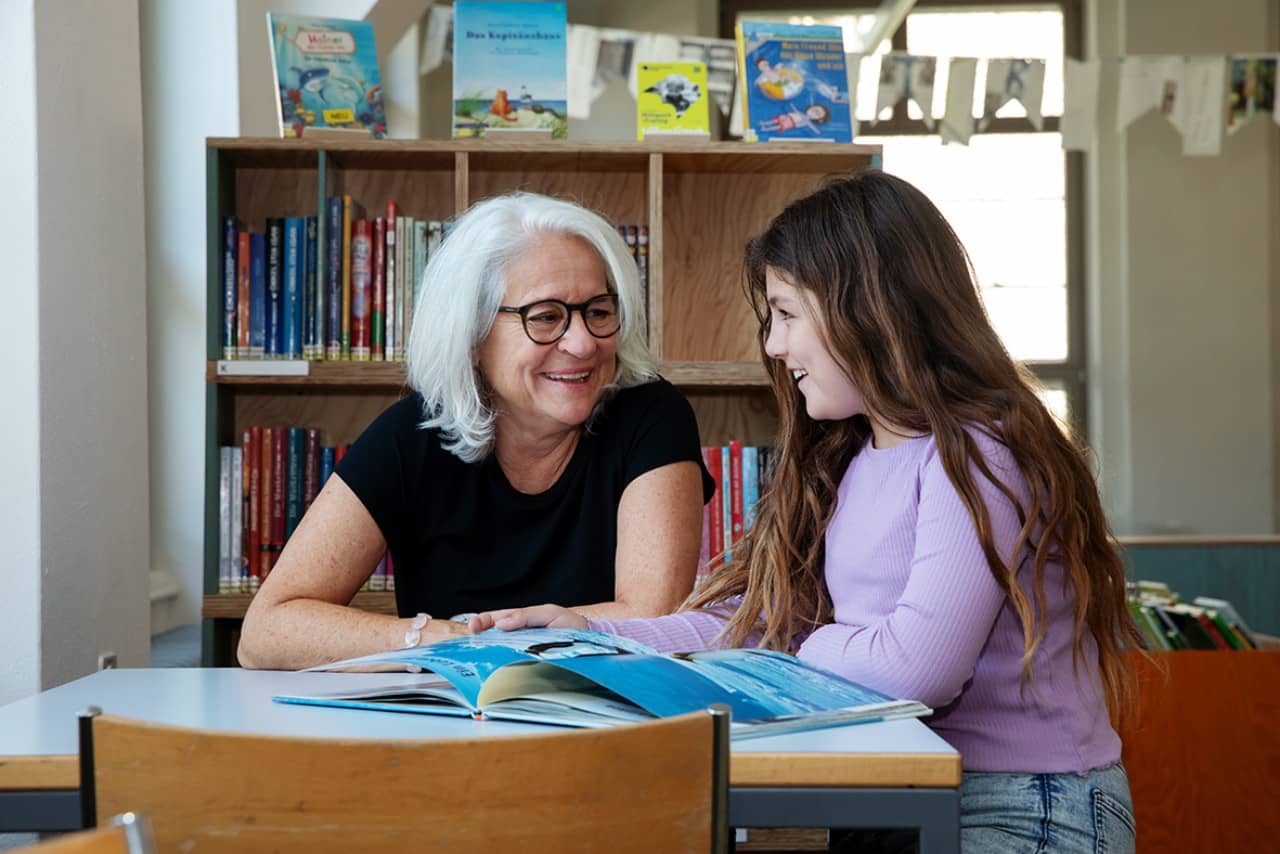 Eine ältere Frau mit grauen, schulterlangen Haaren sitzt mit einem jungen Mädchen an einem Tisch in einer Bibliothek. Sie lesen aus einem Buch und lachen sich an.
