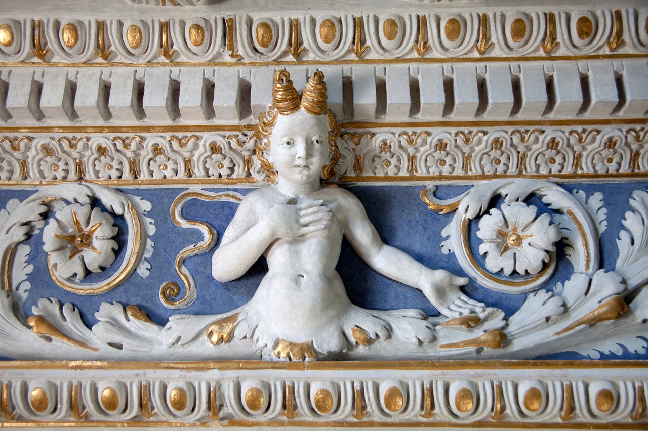 Skulptur einer fantasievollen Figur mit Hörnern, einem menschlichen Oberkörper und einem Fischschwanz, vor blau-goldenem Relief mit floralen Ornamenten.