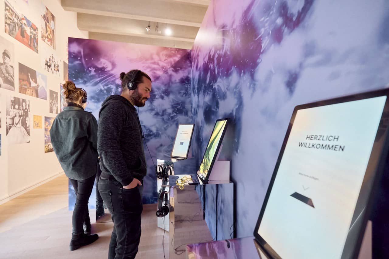 Zwei junge Erwachsene mit Kopfhörern vor einem Display in der Ausstellung