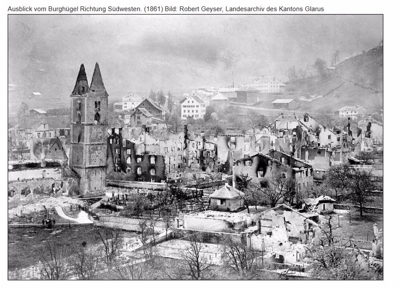 Historische Aufnahme einer zerstörten Stadt mit ausgebrannten Gebäuden und einer Kirche mit beschädigtem Turm inmitten der Ruinen. Schnee bedeckt Teile der Umgebung.