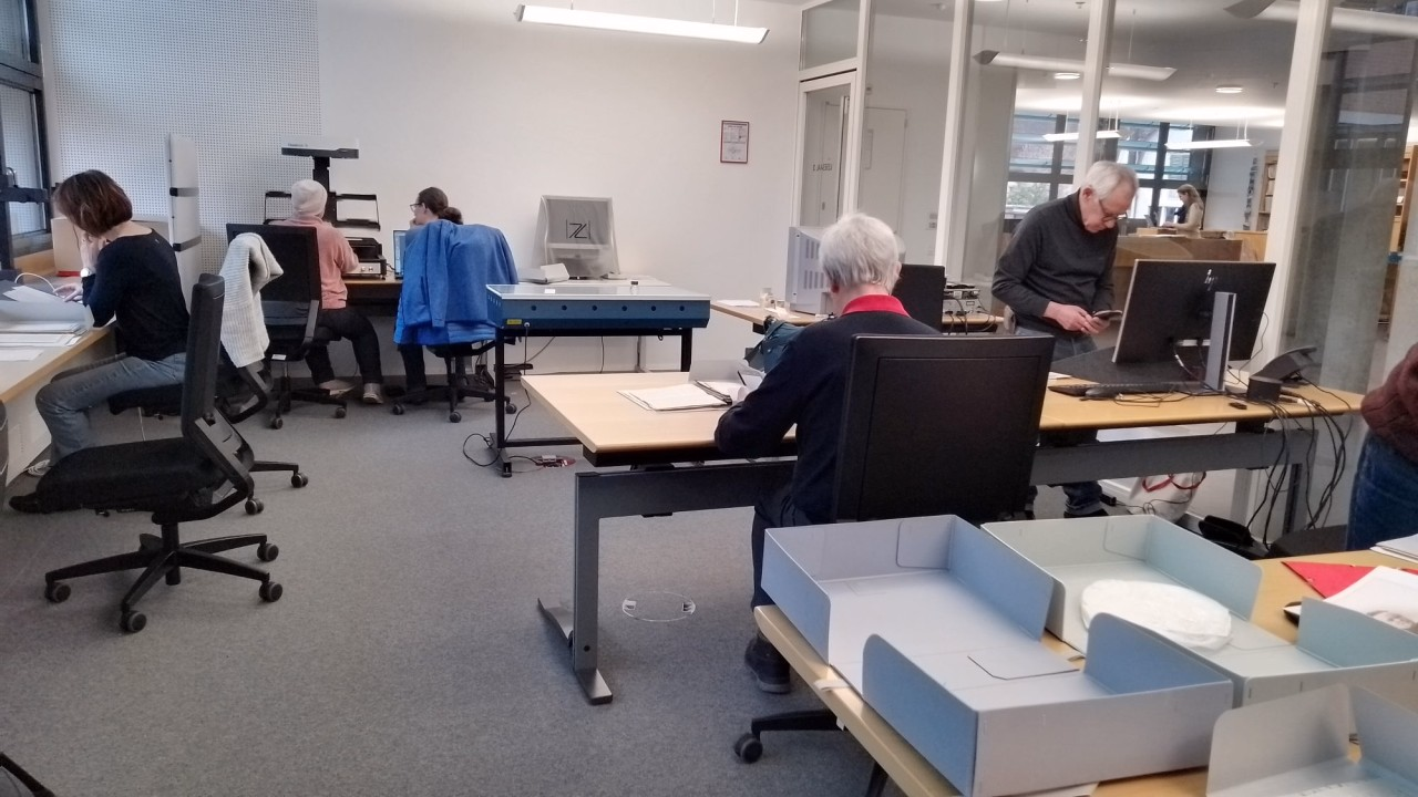 Fünf Personen sitzen verteilt in einem Büro an Tischen mit Archivschachteln und Dokumenten.