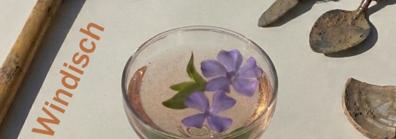 Ein Glas mit rosafarbenem Inhalt und zwei violetten Blumen, daneben alte Werkzeuge auf hellem Hintergrund. Im Bild steht das Wort Windisch.