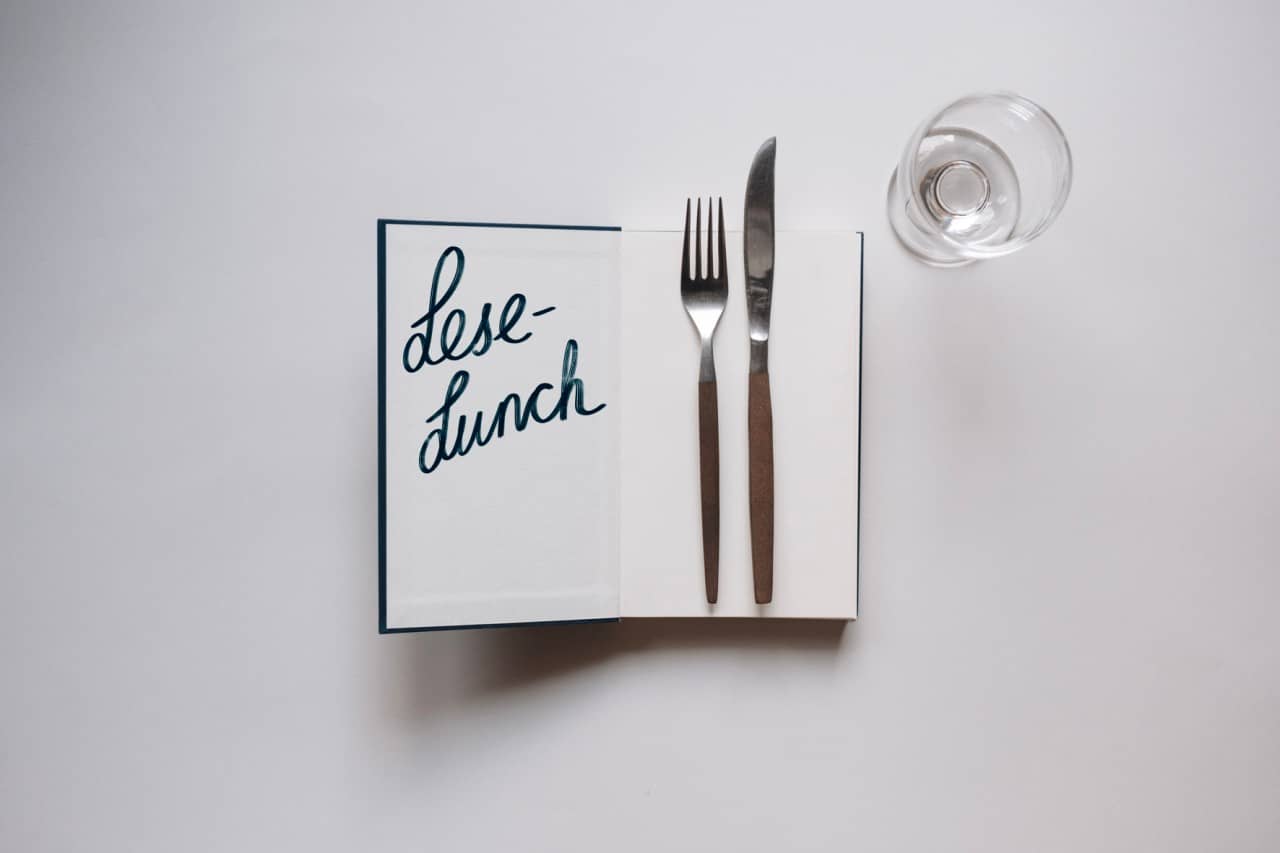 Auf einem weissen Untergrund liegt ein aufgeschlagenes Buch: Auf der Innenseite des Umschlags steht "Lese-Lunch" geschrieben, auf der rechten Seite liegen eine Gabel und ein Messer. Rechts oben nebem dem Buch steht ein leeres Weinglas.