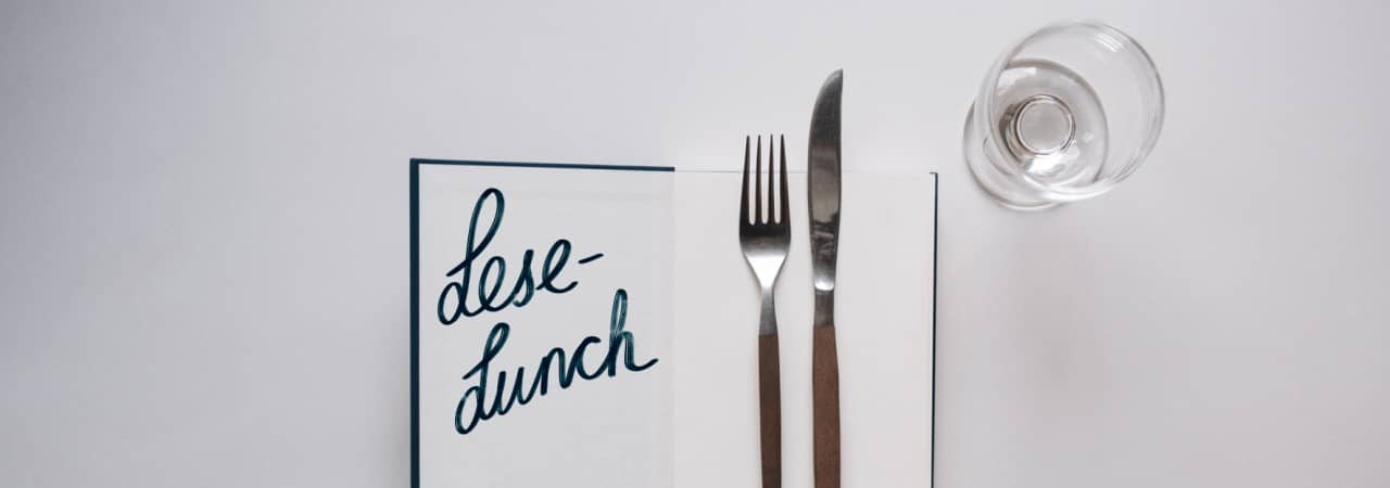 Auf einem weissen Untergrund liegt ein aufgeschlagenes Buch: Auf der Innenseite des Umschlags steht "Lese-Lunch" geschrieben, auf der rechten Seite liegen eine Gabel und ein Messer. Rechts oben nebem dem Buch steht ein leeres Weinglas.