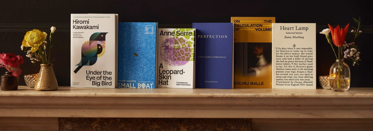 Auf einem Marmorboard sind sechs Buchcover nebeneinander ausgestellt; links und rechts steht jeweils kleine Vasen mit einzelnen Blumen.