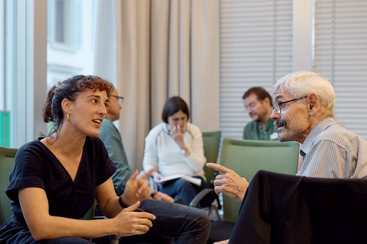 Eine Frau und ein Mann gestikulierend im Gespräch. Im Hintergrund weitere Personen.