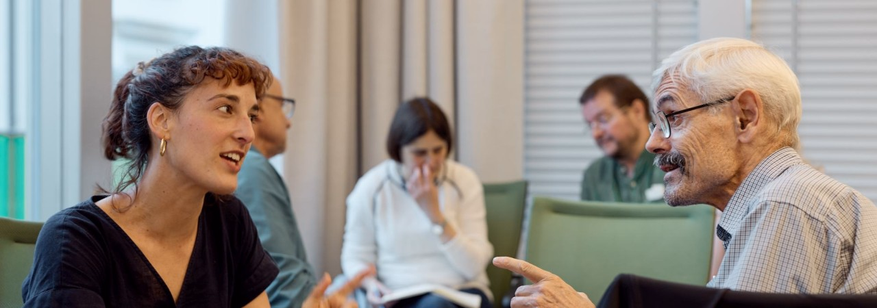 Eine Frau und ein Mann gestikulierend im Gespräch. Im Hintergrund weitere Personen.