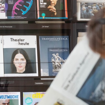 Blick durch Glas auf eine Frau, die vor einem Bücherregal sitzt und eine Zeitung liest.
