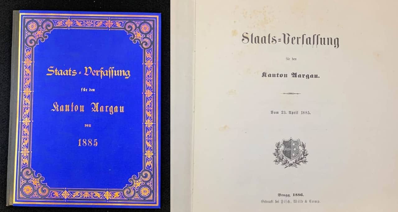 Auf der linken Seite: Staatsverfassung von 1885 mit blauem Einband und goldenem Zierrand. Auf der rechten Seite: Titelblatt Staatsverfassung von 1885 in alter Schrift mit kleinem Wappen auf der unteren Hälfte.