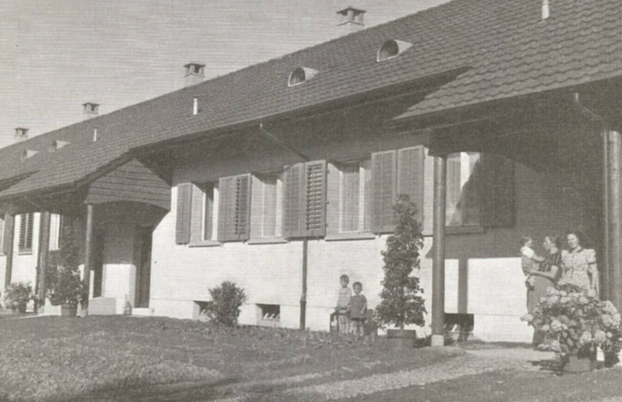 Schwarzweiss-Fotografie – zeigt die der Strasse zugewandten Hauseingänge – Hauseingänge sind überdacht – Frauen und Kinder sind sichtbar – auf dem Dach hat es für jedes Haus einen Kamin (Holzheizung)