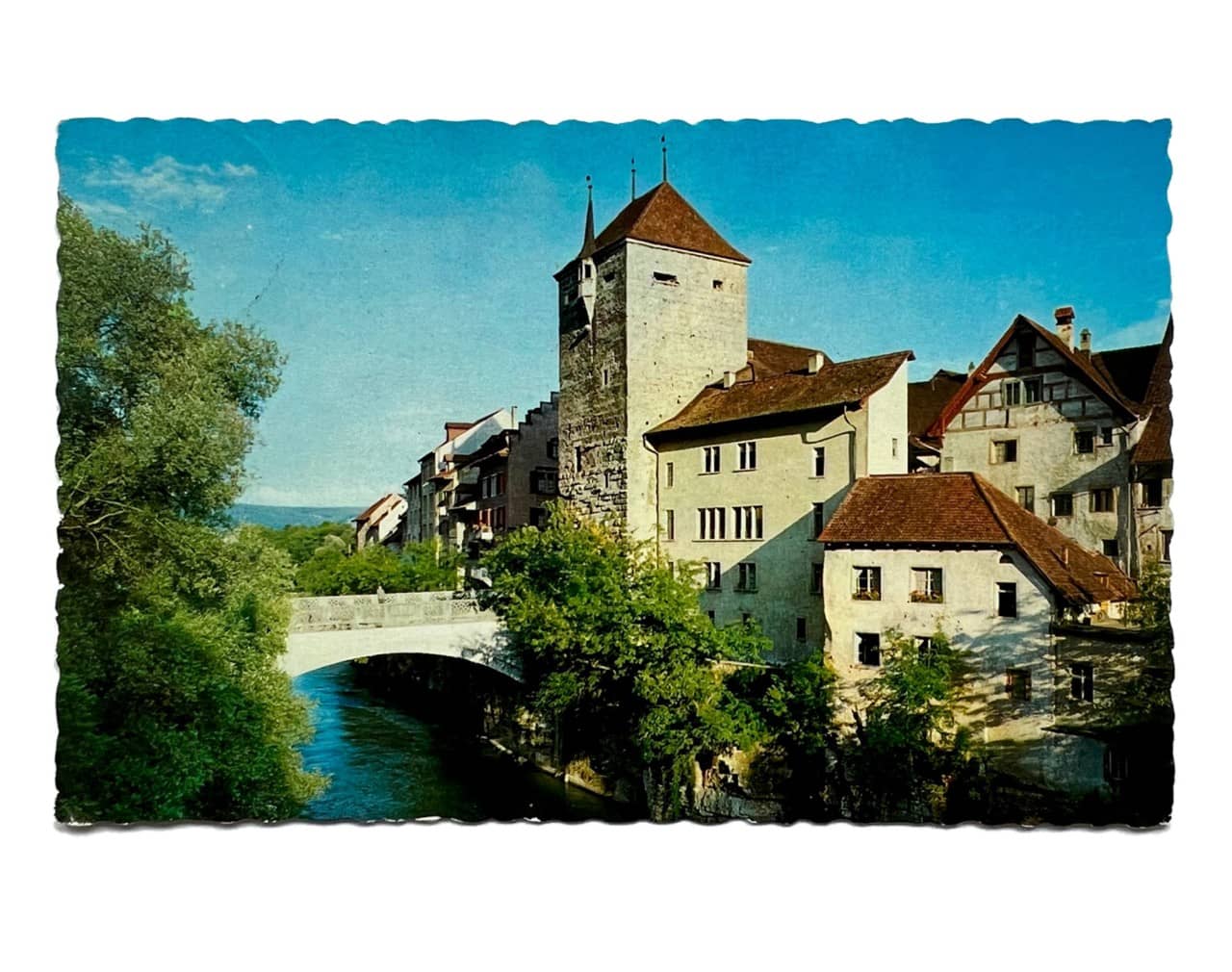 Eine Postkarte mit gewellten Rändern. Darauf zu sehen, ist ein Farbfoto von der Aarebrücke in Brugg. Auf der rechten Seite ragt der schwarze Turm in die höhe, links daneben breitet sich die Brugger Altstadt aus.