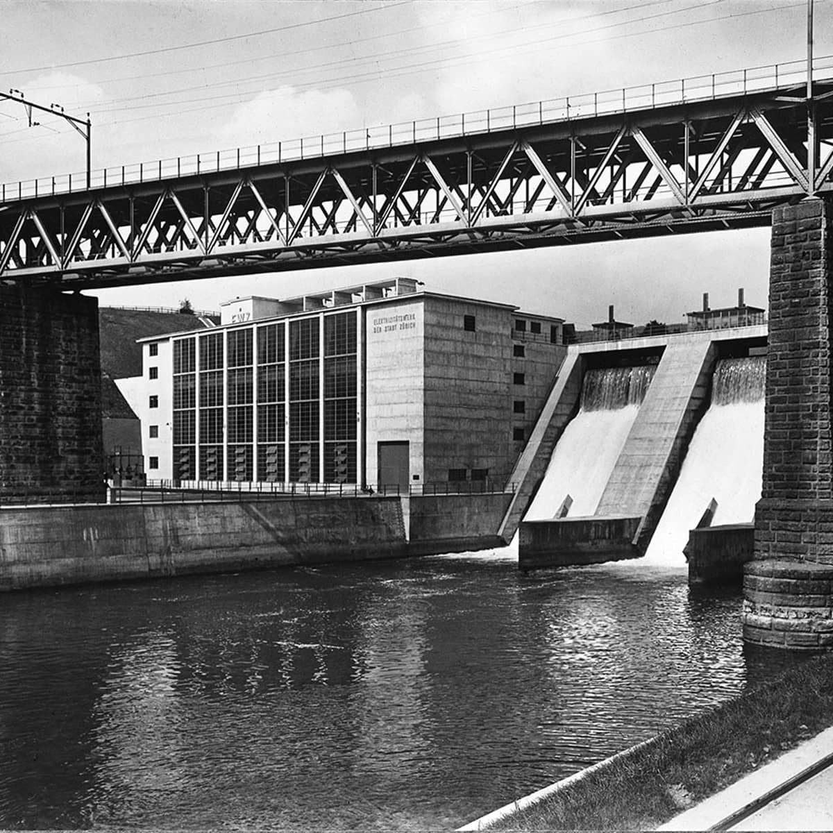 Eine schwarz-weiss Fotografie des ewz Wettingen mit Blick auf die Staumauer und die Bahngleise, die oben drüber ziehen.