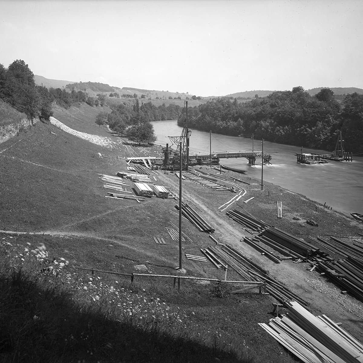 Eine Schwarzweissfotografie am Ufer der Limmat: Es liegen Holzlamellen auf der Wiese, in der Limmat ragen bereits einige Konstruktionen aus dem Wasser.