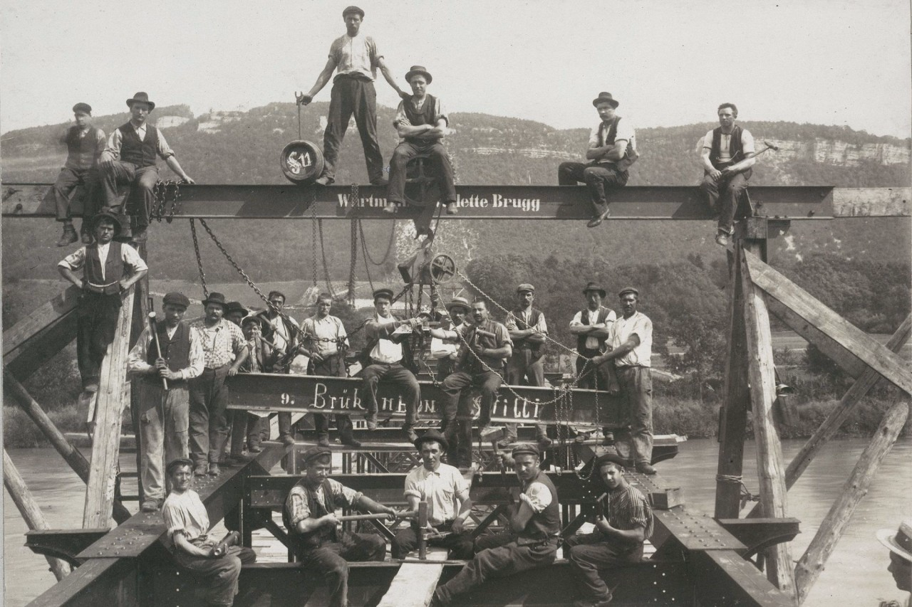 Schwarz-weiss-Foto von Arbeitern sitzend und stehend auf einer Brückenkonstruktion.