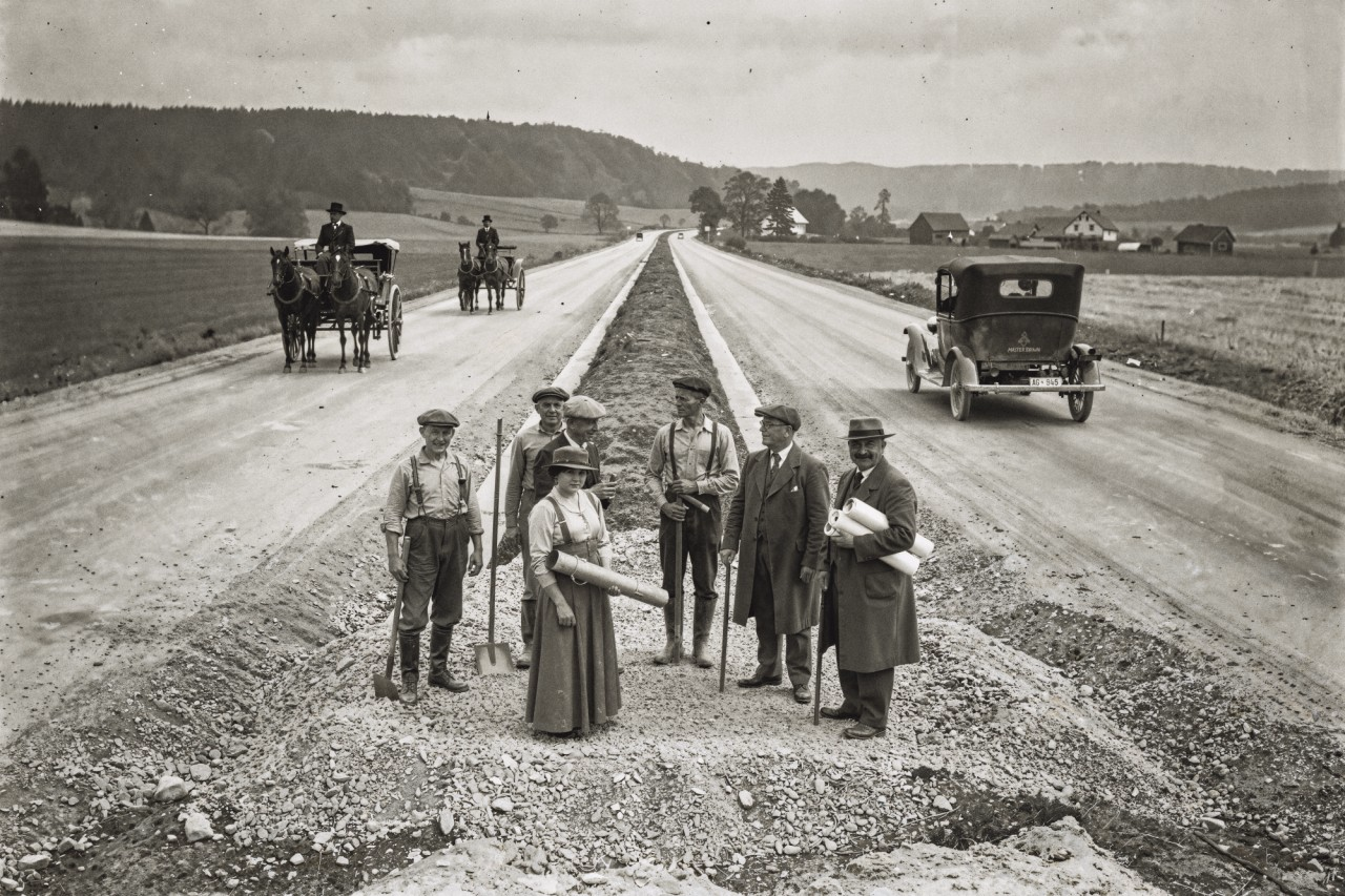 Schwarzweiss-Fotografie, die den Bau einer ersten Autobahn zeigt, auf der sowohl Pferdekutschen als auch Autos fahren. Im Vordergrund eine Gruppe Arbeiter und eine einzelne Frau, die mit Bauplänen und Schaufeln posieren.