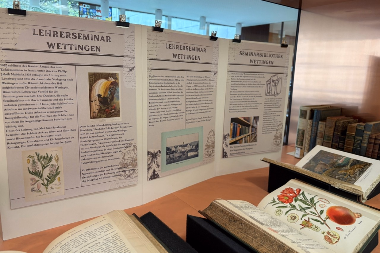 Blick schräg in eine geöffnete Vitrine auf gestaltete Stellwand mit Text und Bild und aufgeschlagene, farbig illustrierte Bücher.