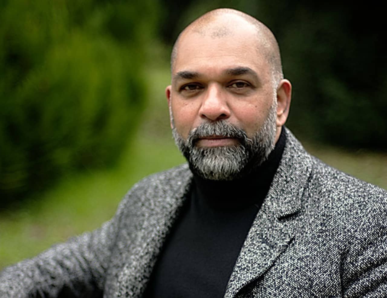 Eine Portraitfotografie des Autors Sunil Mann. Der Hintergrund ist grasgrün und verschwommen. Der Autor ist glatzköpfig, trägt einen grauen Bart und einen grauen Blazer.
