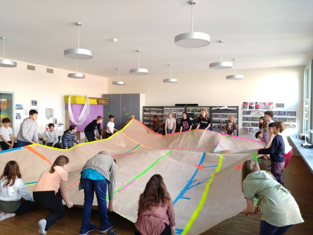 Eine Schulklasse hält ein riesiges, zusammengeklebtes Papier im Schulzimmer