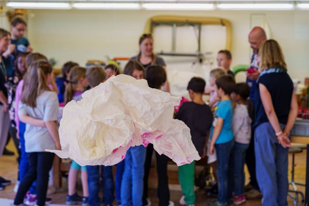 Ein Obekt aus Papier hängt an einem Faden. Im Hintergrund sind Schülerinnen und Schüler sowie fünf Erwachsene Personen.