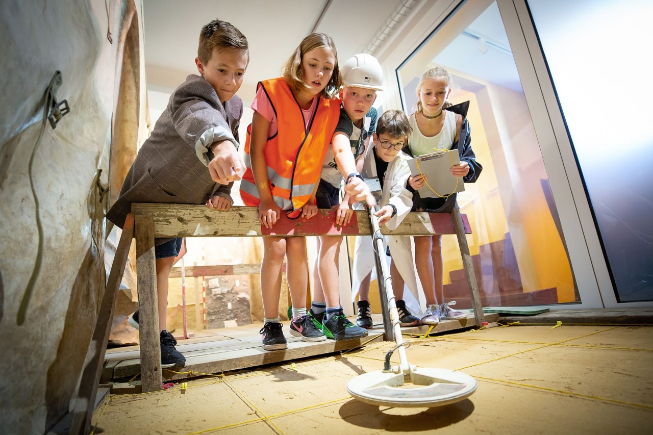 Vier Schülerinnen und Schüler untersuchen mit einem Metalldetektor eine abgesteckte Fläche. 