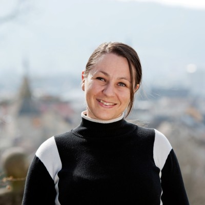 Eine lächelnde Frau vor einer Stadtlandschaft mit unscharfen Gebäuden und Bergen im Hintergrund.