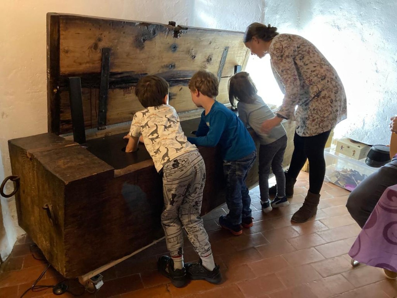 Drei Kinder strecken ihren Arm durch Löcher in eine grosse Holzkiste, daneben steht eine erwachsene Person.