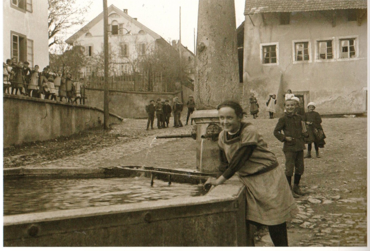 Auf einer historischen Fotografie befinden sich mehrere Kinder und Jugendliche. Ein Mädchen fasst in einen Brunnen. 