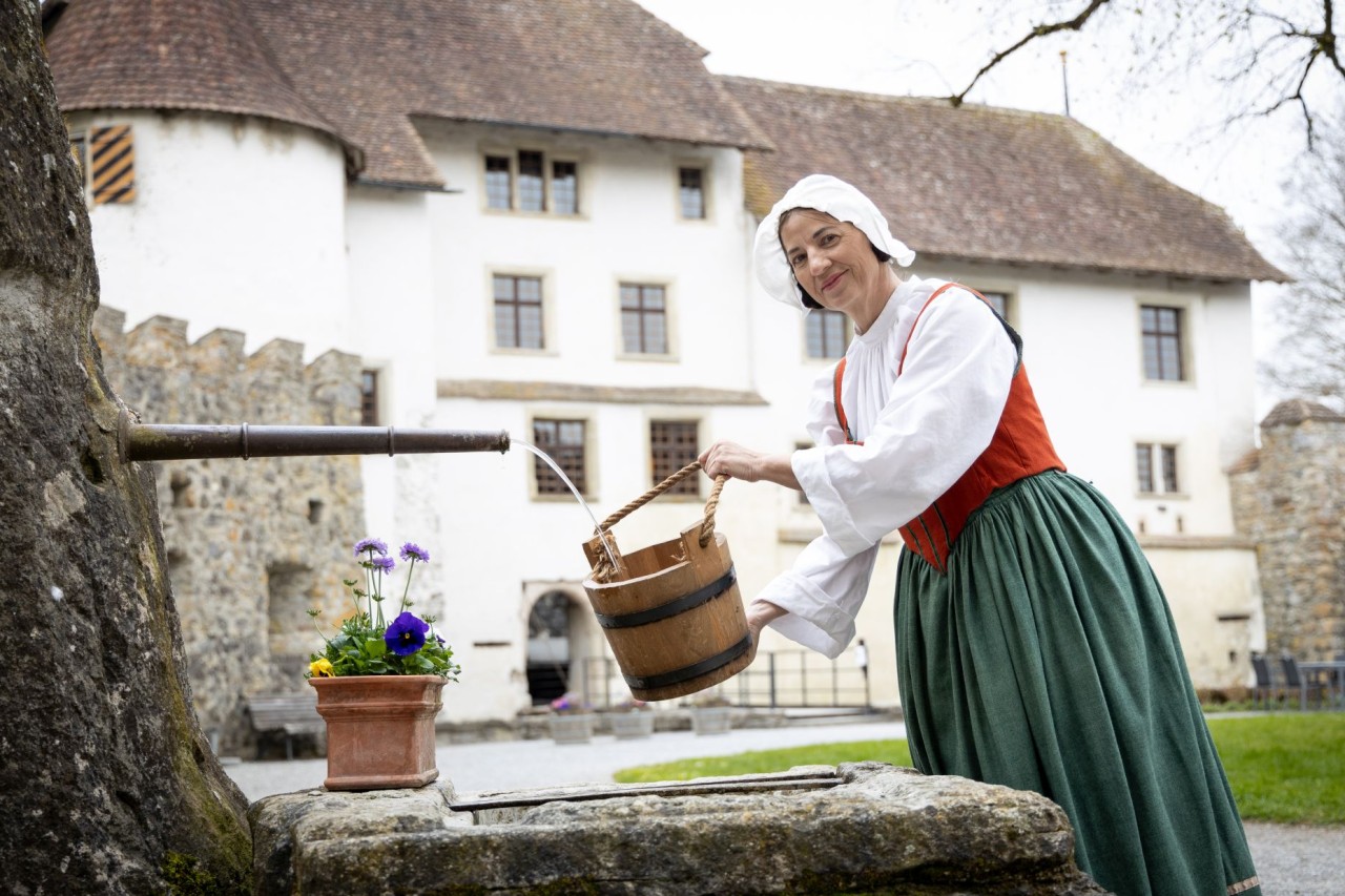 Person in historischer Kleidung füllt draussen vor einem Schloss einen Holzeimer an einem Brunnen.