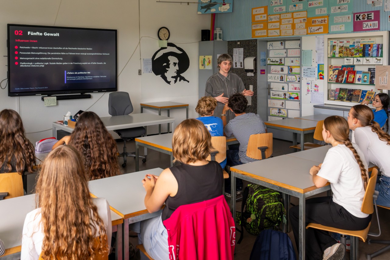 Eine Gruppe von Schülerinnen und Schülern sitzt in einem Klassenzimmer, eine Frau steht vorne und hält eine Präsentation. Auf dem Bildschirm ist der Titel "Fünfte Gewalt" sichtbar.