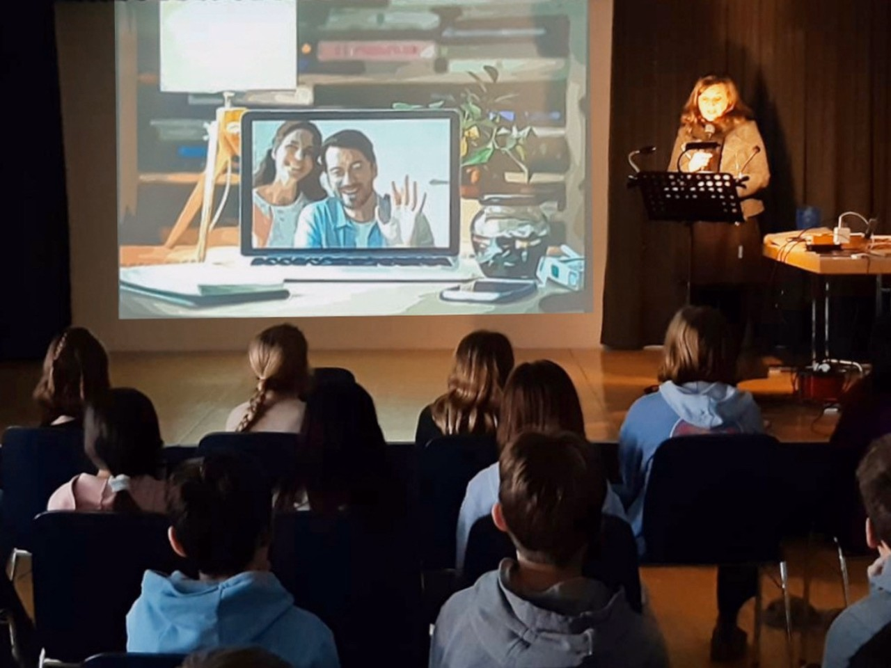 Schülerinnen und Schüler sitzen auf Stühlen vor einer Leinwand, die ein Laptop in einem Büro mit zwei winkenden Personen auf dem Bildschirm zeigt. Neben der Leinwand steht eine Frau an einem Rednerpult.