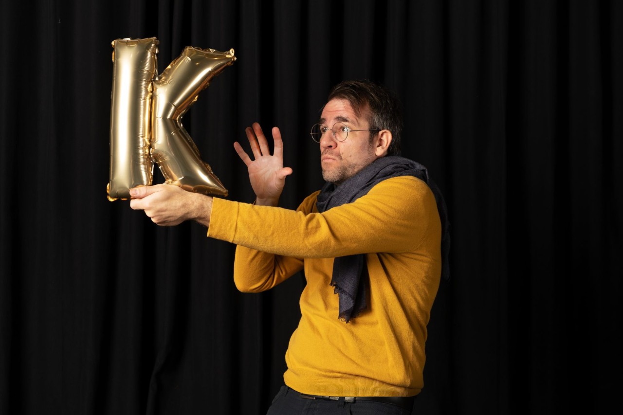 Person in gelbem Pulli hält einen goldenen Ballon in Form des Buchstabens K vor schwarzem Hintergrund.