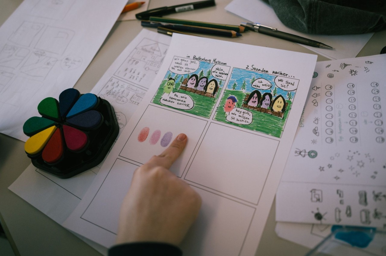 Hand gestaltet einen Comicstreifen mit Fingerabdrücken und bunten Zeichnungen auf einem Tisch mit Stiften und Papier.