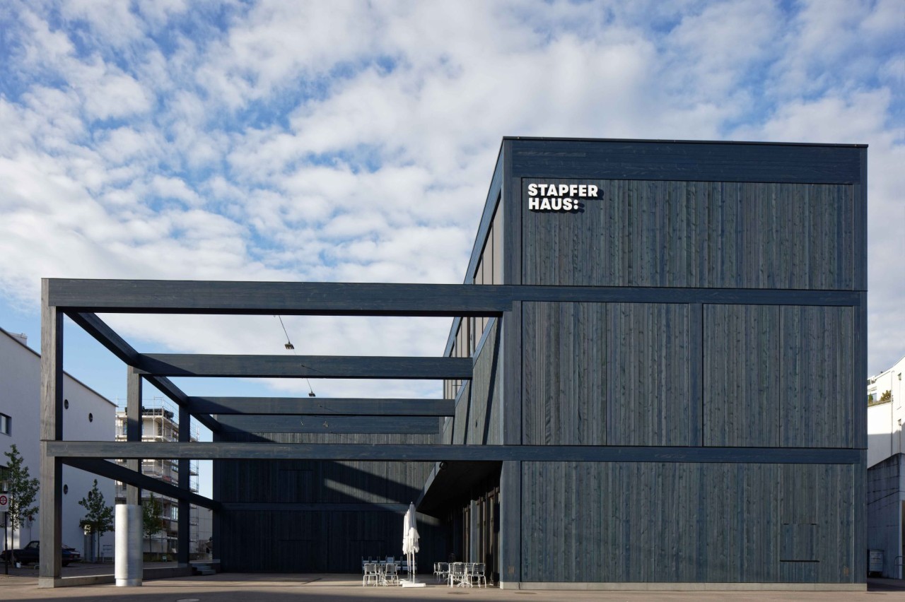Ein modernes Gebäude mit dunkler Holzfassade, breiten rechteckigen Metallstrukturen und der Aufschrift Stapferhaus. Wolken am Himmel.