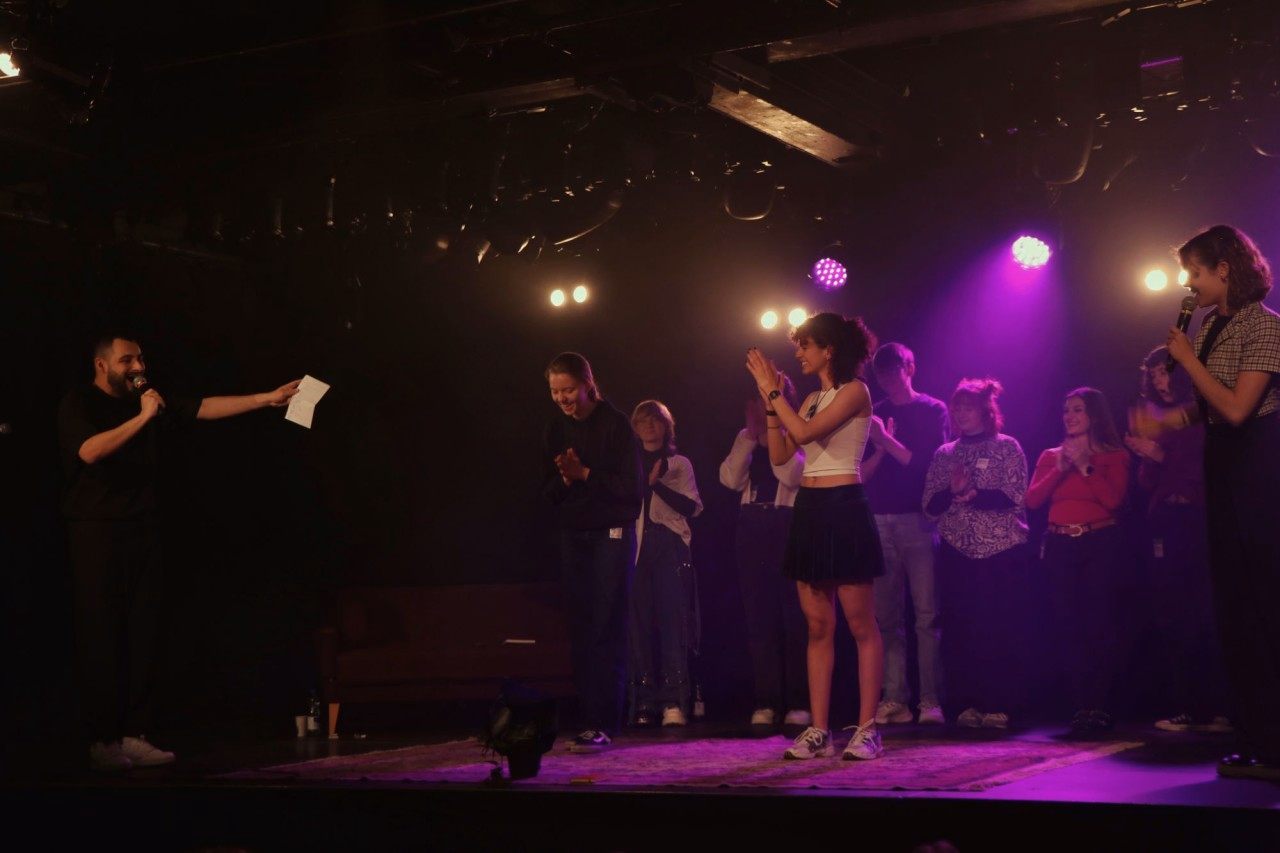 Gruppe junger Menschen steht auf einer Bühne im Scheinwerferlicht und applaudiert während einer Moderation.