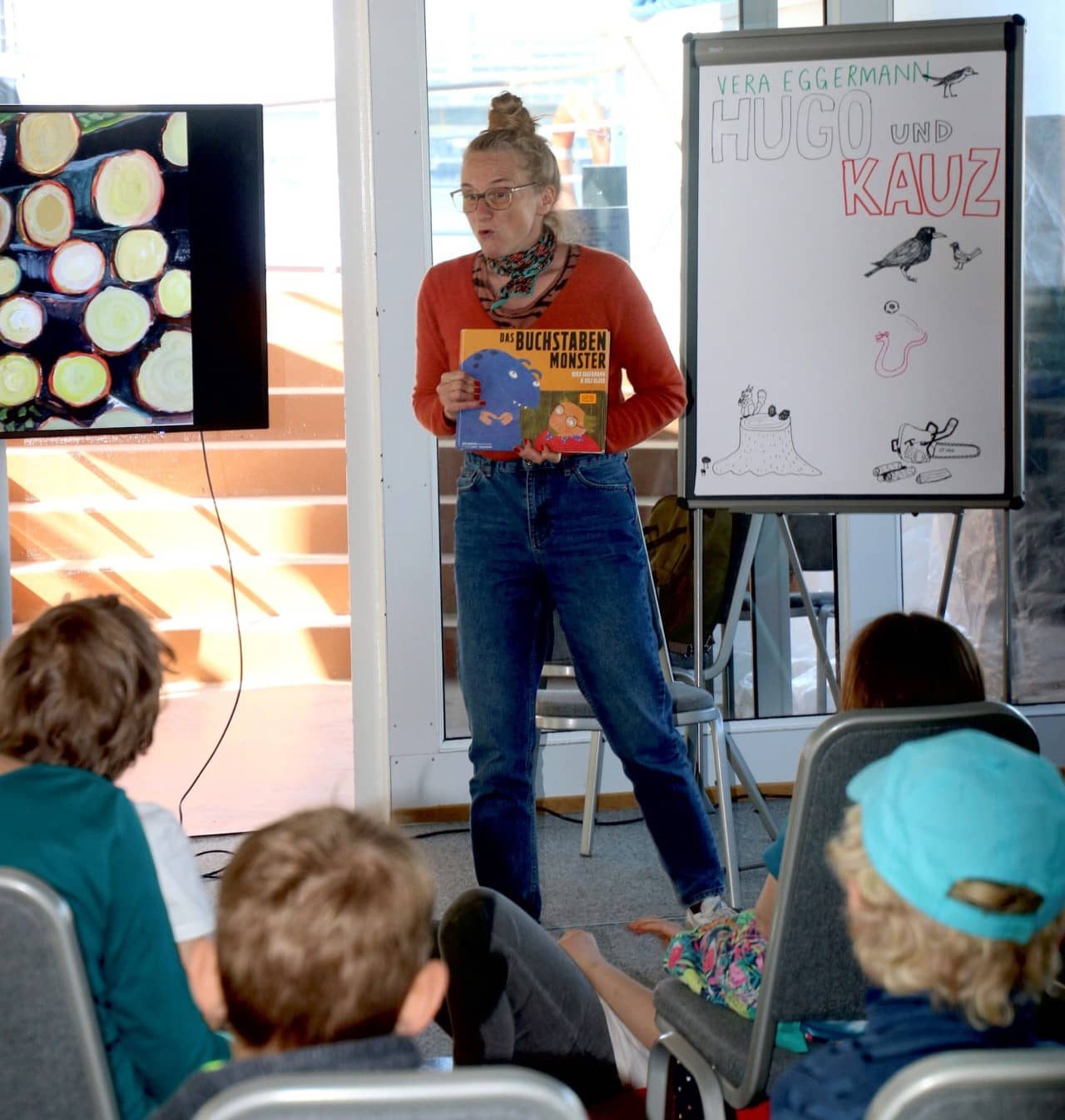 Eine Frau hält ein Buch und spricht zu einer Schulklasse. Im Hintergrund befindet sich ein Flipchart mit Illustrationen.