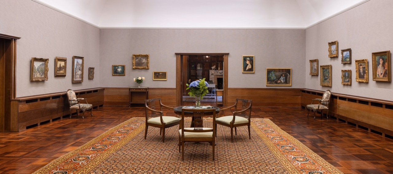 Innenraum mit Tisch, Stühlen und opulentem Teppich in der Mitte und an den Wänden Gemälde.